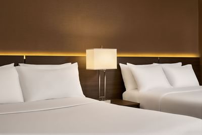Double guest room with white linens, a nightstand with a lit lamp, and a wall with a lit panel above the headboards.