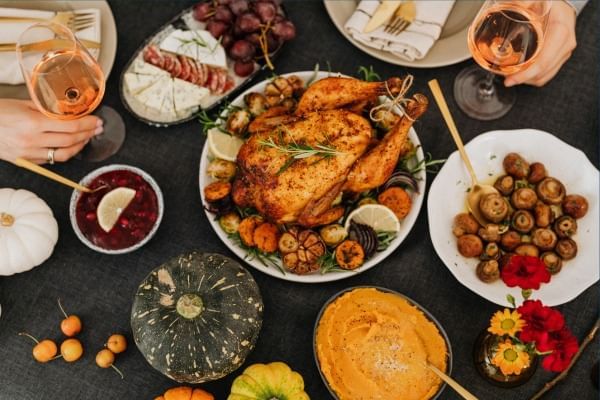 Hands holding wine glasses over a table with roasted turkey, sides, and seasonal decor.