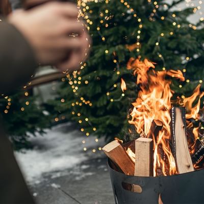 Hand fügt Holz zu einem Feuer in einem Metallkorb neben einem beleuchteten Weihnachtsbaum hinzu.