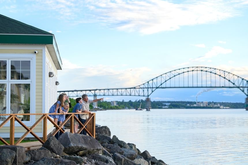 Family on a wooden deck by a yellow building under a blue sky, with a massive arch bridge at Rodd Miramichi River