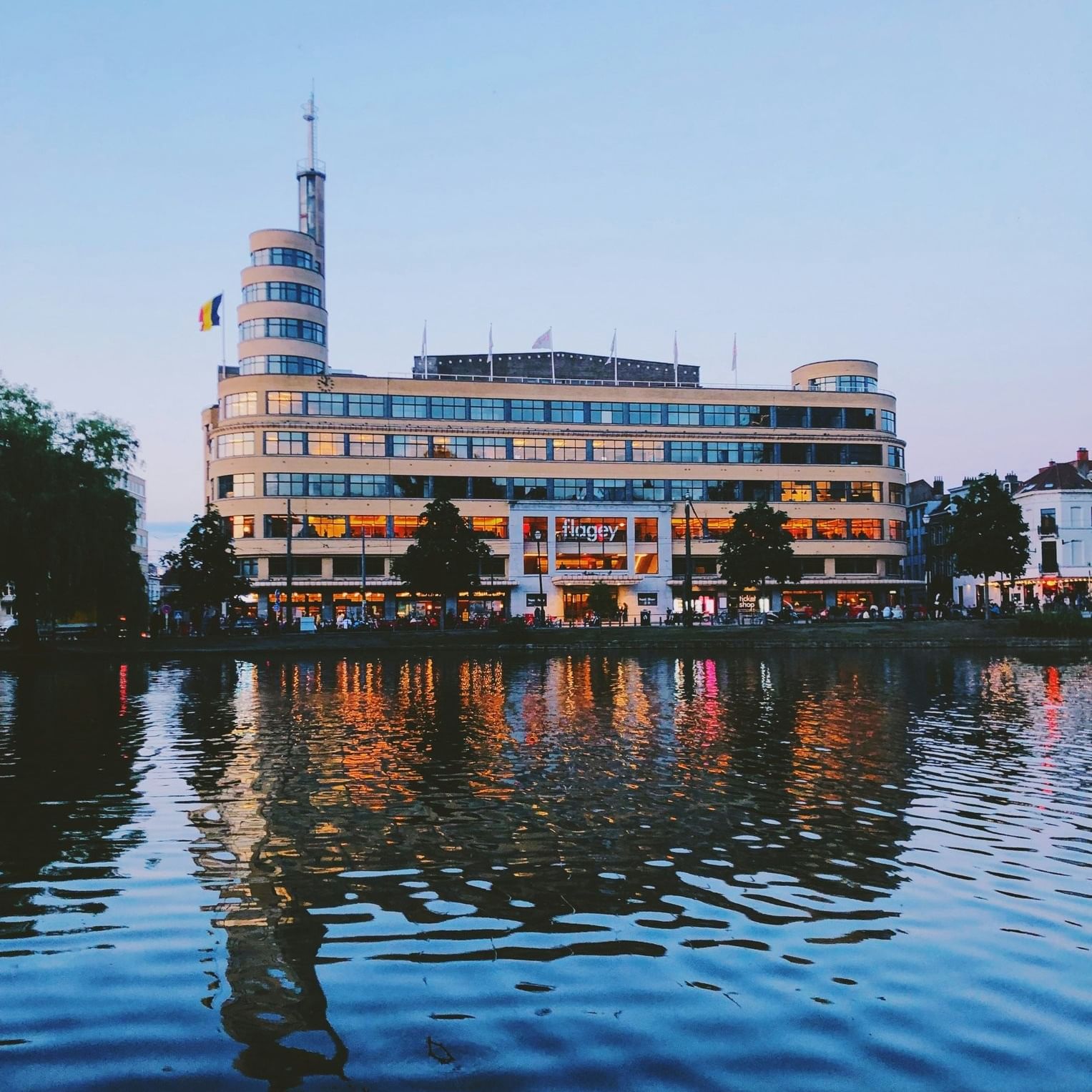 Flagey building with glowing windows by a calm lake reflecting city lights near Hotel Barsey by Warwick