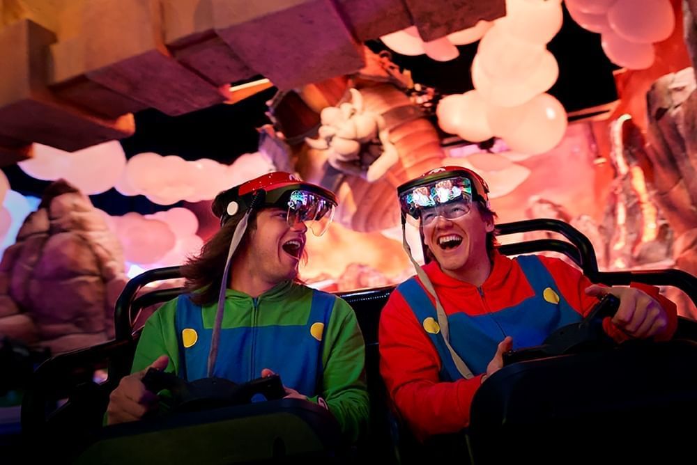 Two men in Mario and Luigi sweatshirts smile as they drive a cart beneath bubble clouds. 