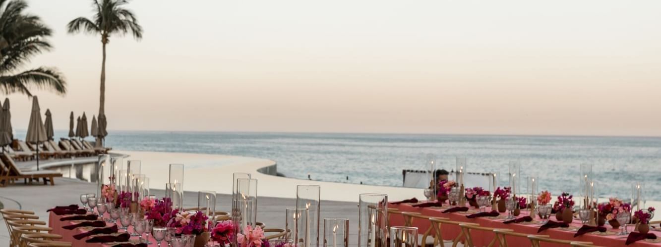 Long dining tables with pink linens and floral centerpieces set for an event by the ocean at Marquis Los Cabos Resort