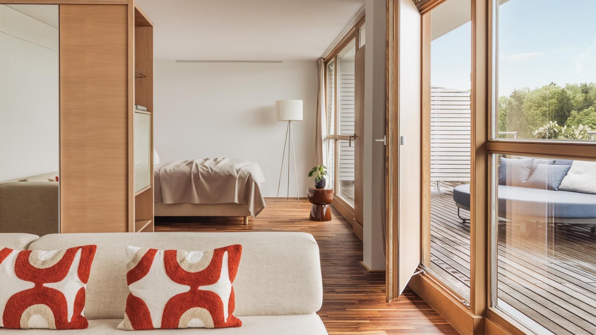 Modernes Zimmer mit Holzboden, Sofa, Bett, Balkon und Ausblick auf Bäume.