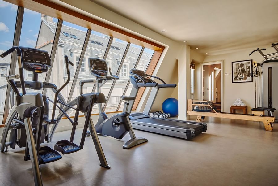 Fitness center with equipment arranged by floor-to-ceiling sloped windows overlooking the city at Hotel Westminster Paris