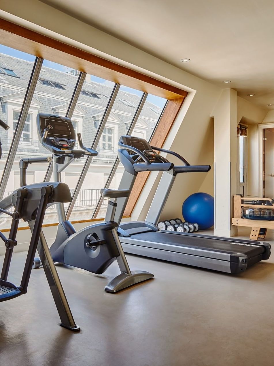 Fitness center with equipment arranged by floor-to-ceiling sloped windows overlooking the city at Hotel Westminster Paris