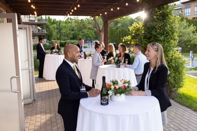 Guests mingling at an outdoor event with tables, flowers, and wine, under string lights.