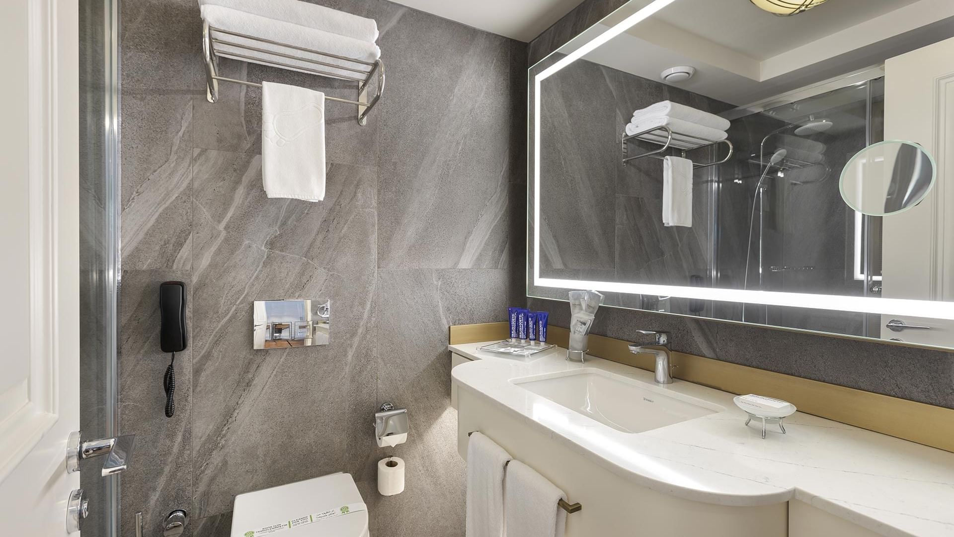 A modern bathroom with gray walls, a white toilet, an illuminated mirror, a sink, towels, and a glass shower enclosure in Executive Room at Titanic Port Bakırköy