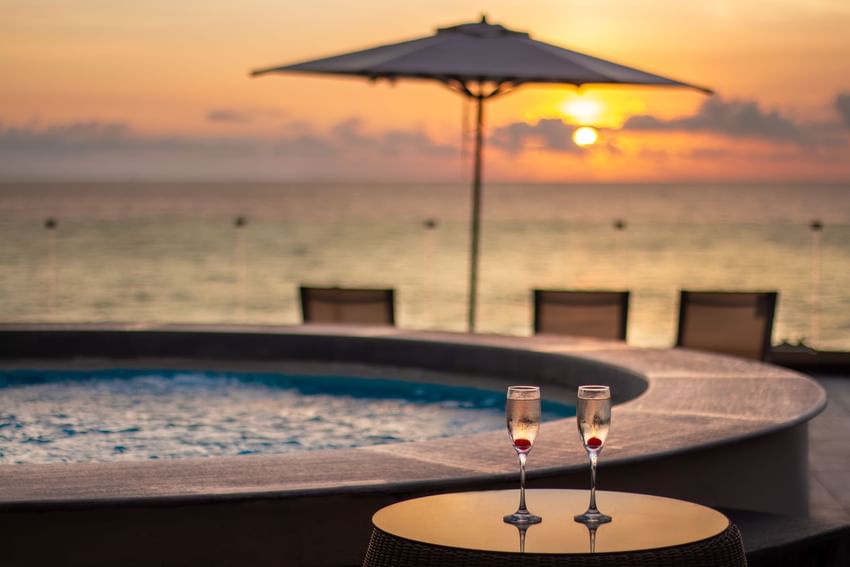 Two champagne glasses with cherries sit on table beside hot tub, overlooking sunset over ocean at Sunset Plaza Beach Resort
