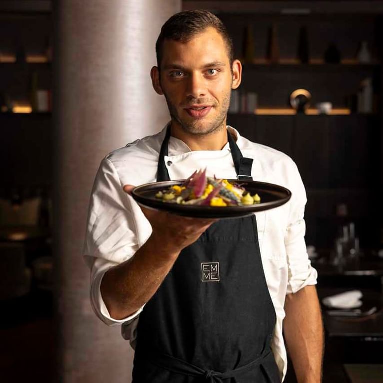 A chef in a white shirt and brown aprons holding a dish in EMME Restaurant at Margutta 19