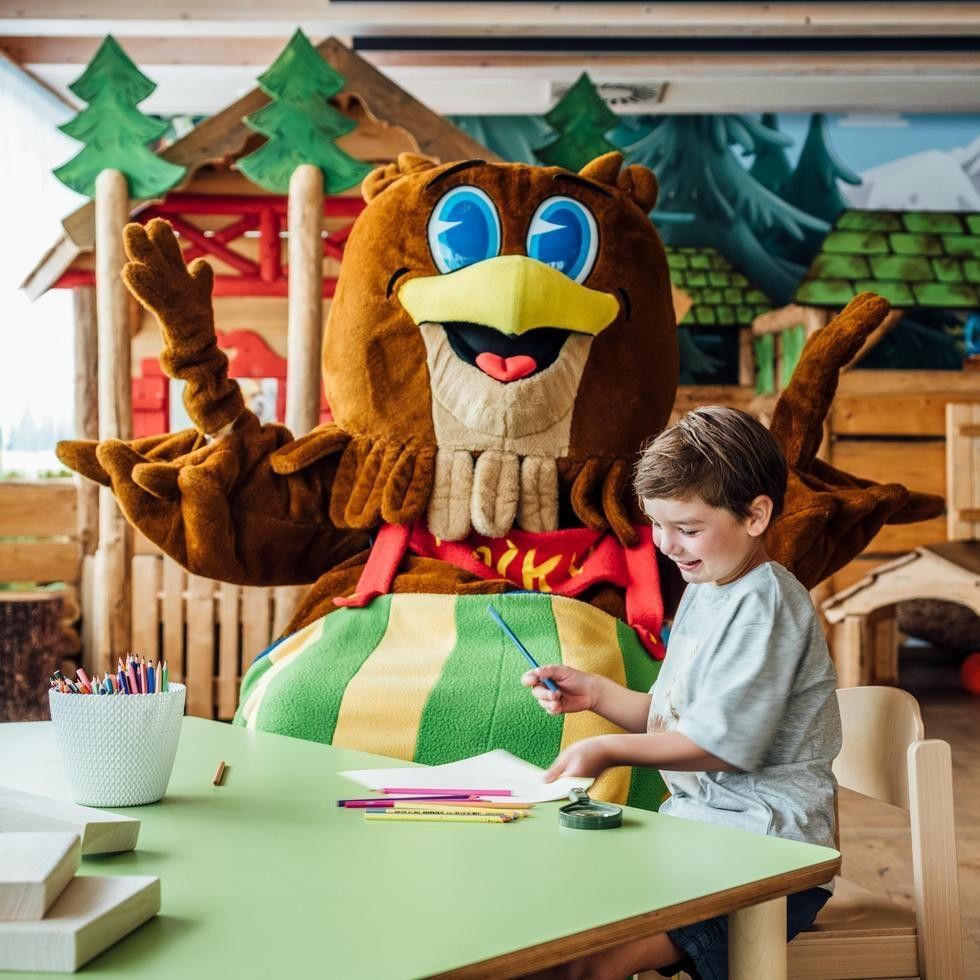 Falkys Weihnachtsakademie mit Maskottchen und Kind beim Basteln an einem Tisch mit Weihnachtsdekoration im Hintergrund.