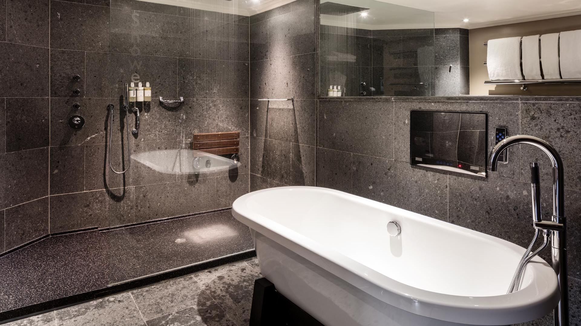 Modern bathroom featuring a freestanding white tub, rainfall shower, and dark stone tiles, creating a sleek, serene space
