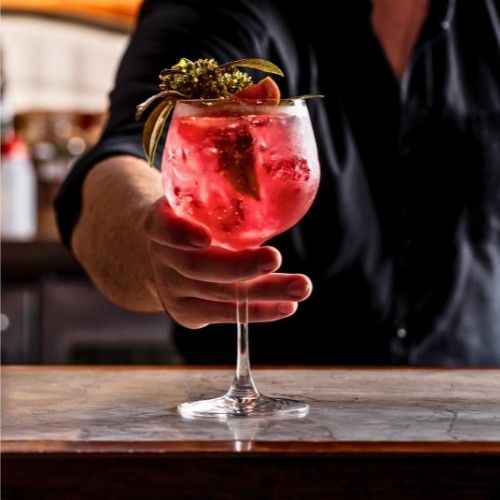 A hand holds a large, stemmed glass filled with a red gin and tonic. The drink is garnished with a orange slice and green leaves. The person's arm and black shirt are visible, as is the marble countertop of a bar.