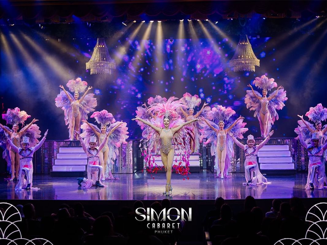Simon Cabaret performers in sparkling feathered costumes pose under dramatic stage lighting during a vibrant dance performance in Patong Beach, Phuket.