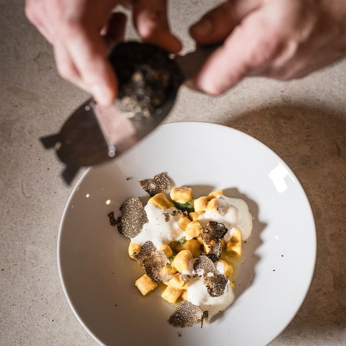 Hände geben schwarzes Essen auf einen weißen Teller mit gewürfelten Kartoffeln, Sahne und schwarzen Pilzen.