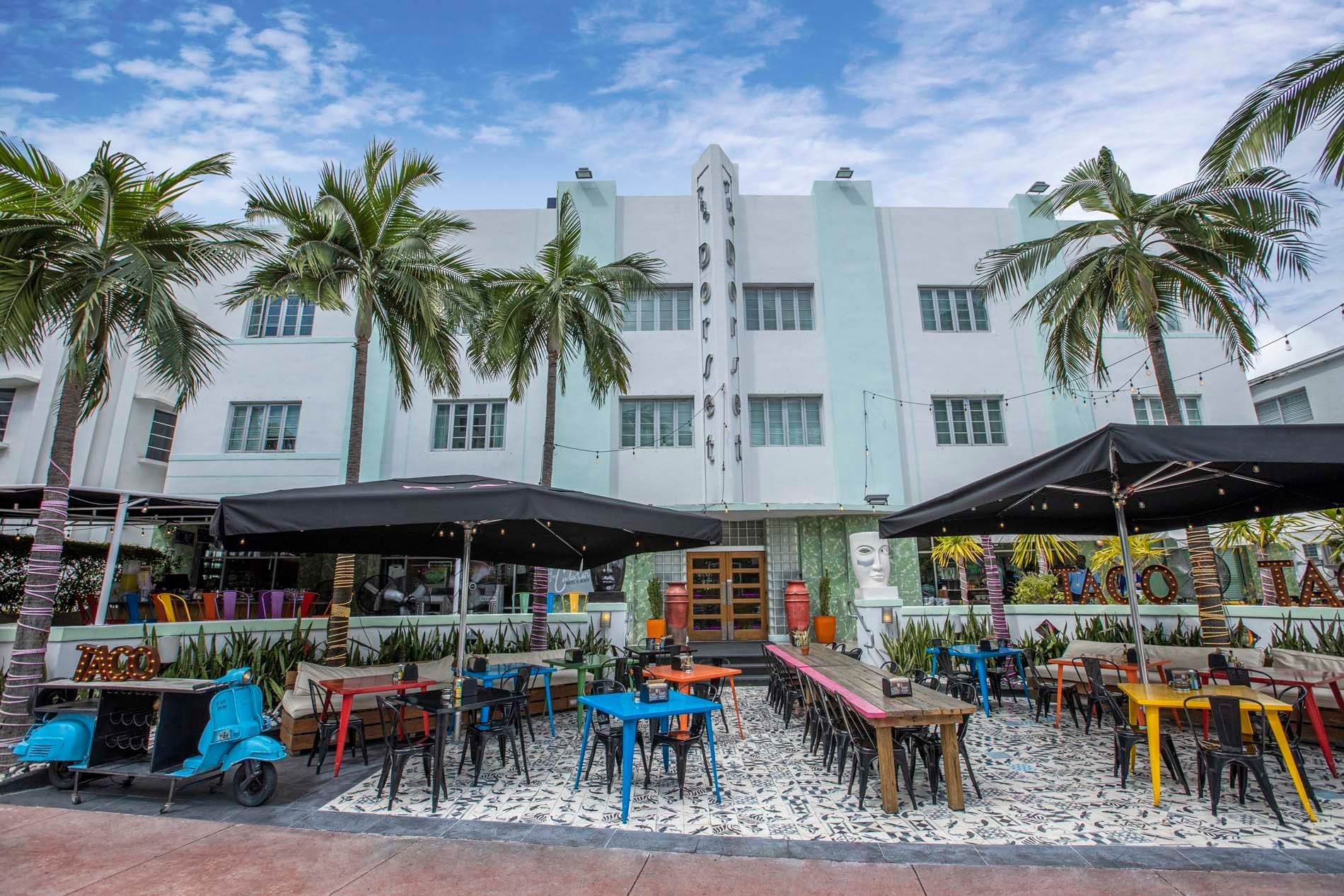 A white building with a patio area featuring colorful tables and chairs, umbrellas, and palm trees.