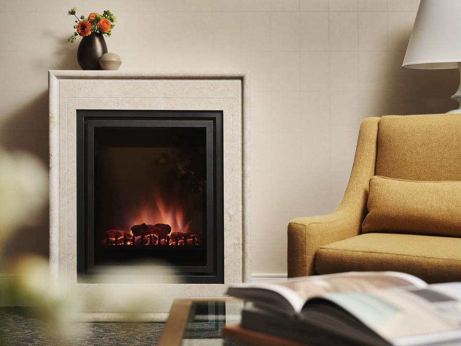 A lit electric fireplace in a room with a chair, lamp, vase, and open book on a table.