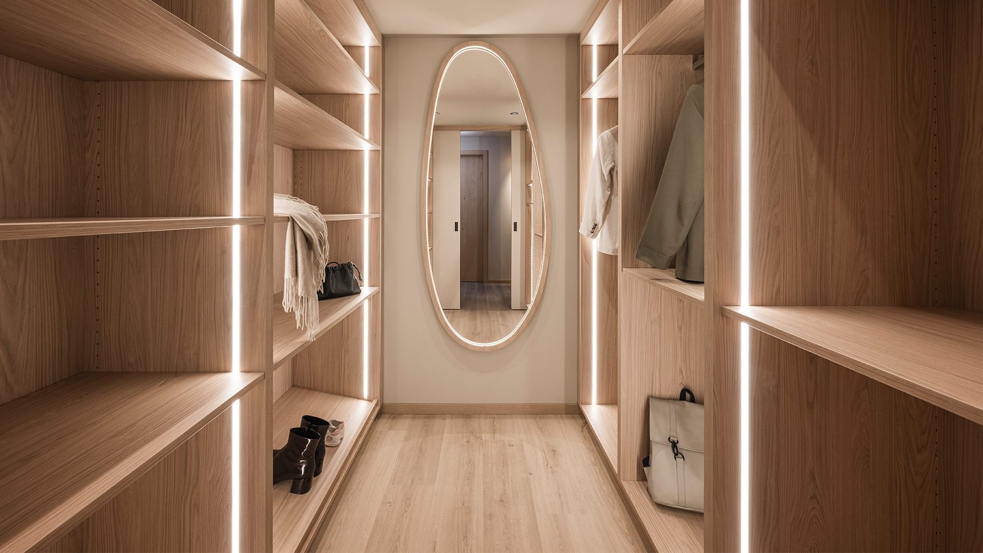 Modernes Ankleidezimmer mit Holzschränken, hellem Boden, beleuchteten Regalen und ovalem Spiegel.