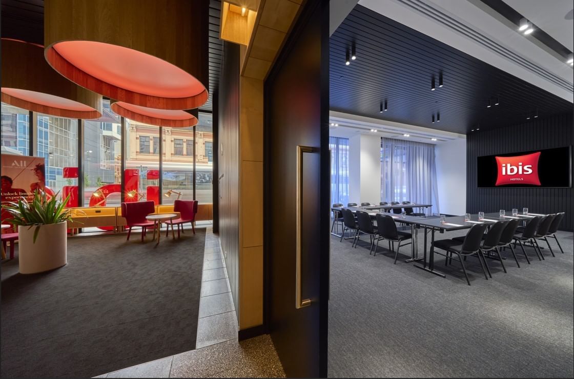 U-shaped table setup facing a wall-mounted TV in The Engine Room, alongside a cozy lounge area at Ibis Adelaide