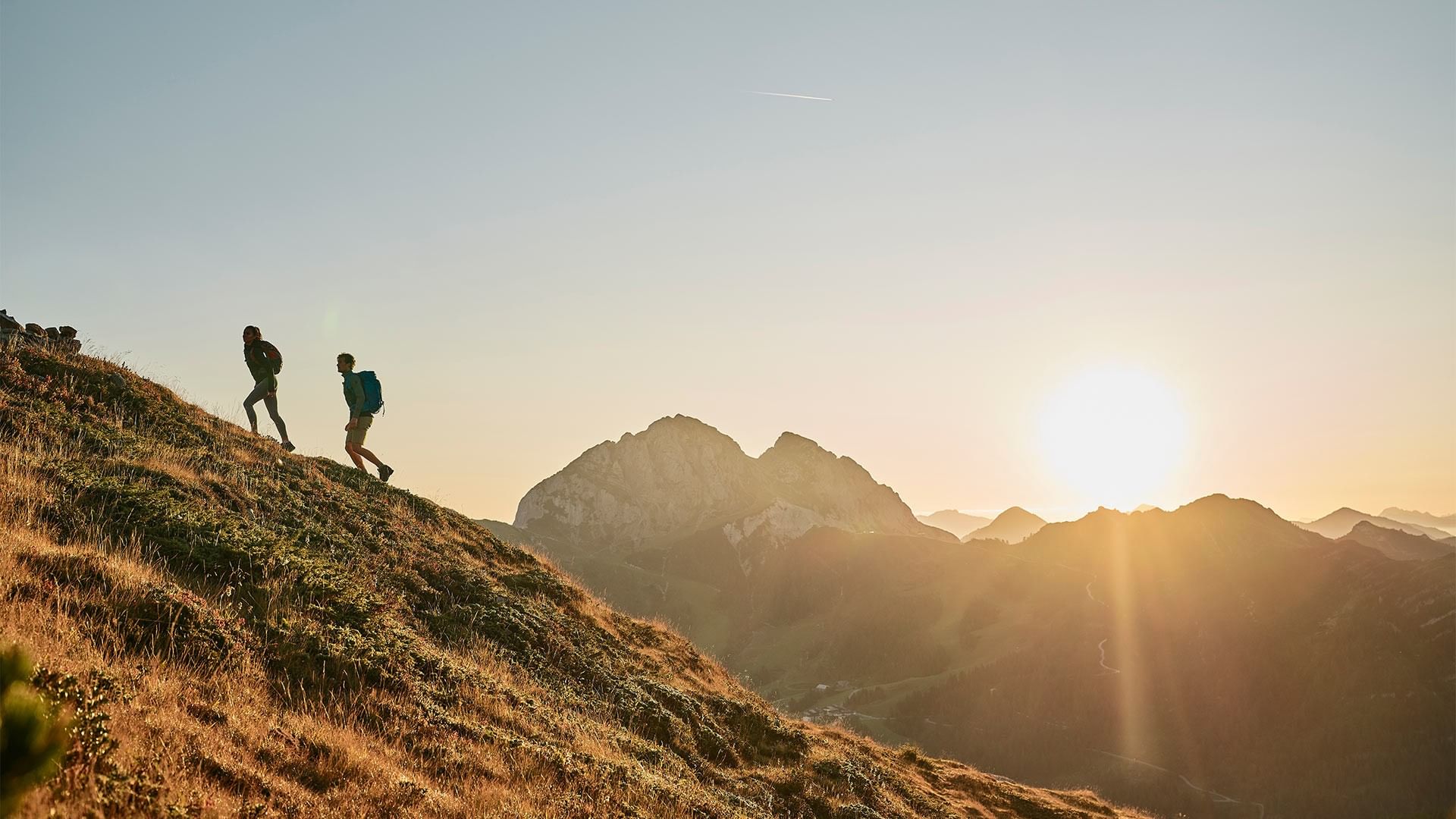 Two hikers with backpacks ascend a grassy mountain slope at sunrise, with a stunning mountain range in the background.
