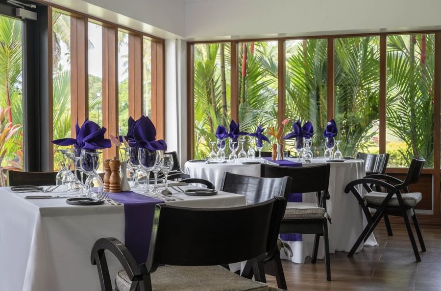 Dining tables with purple napkins by large windows under a white ceiling in Talei Restaurant at The Naviti Resort - Fiji