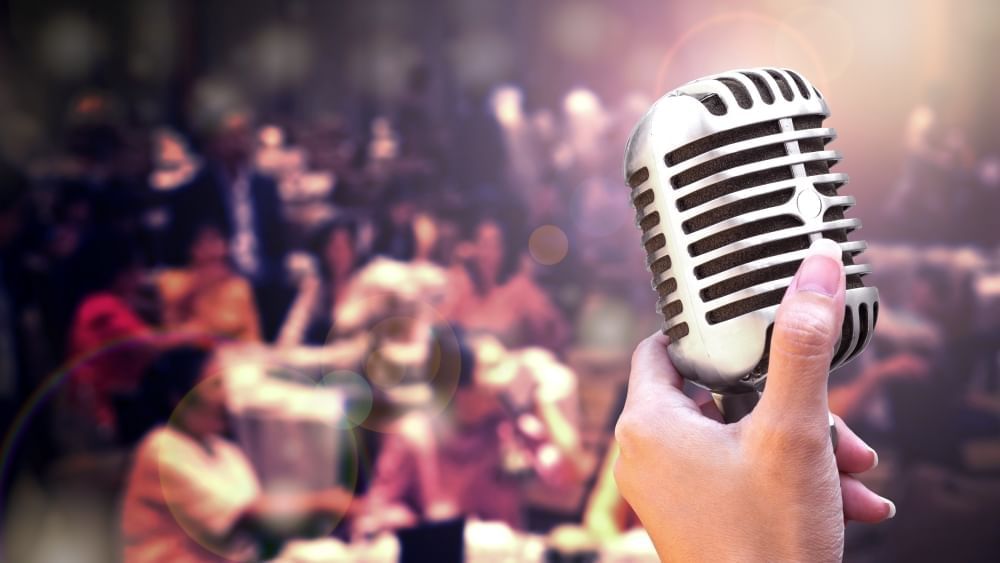 A close-up of a hand holding an old-fashioned microphone looking out over a crowd of out-of-focus diners.