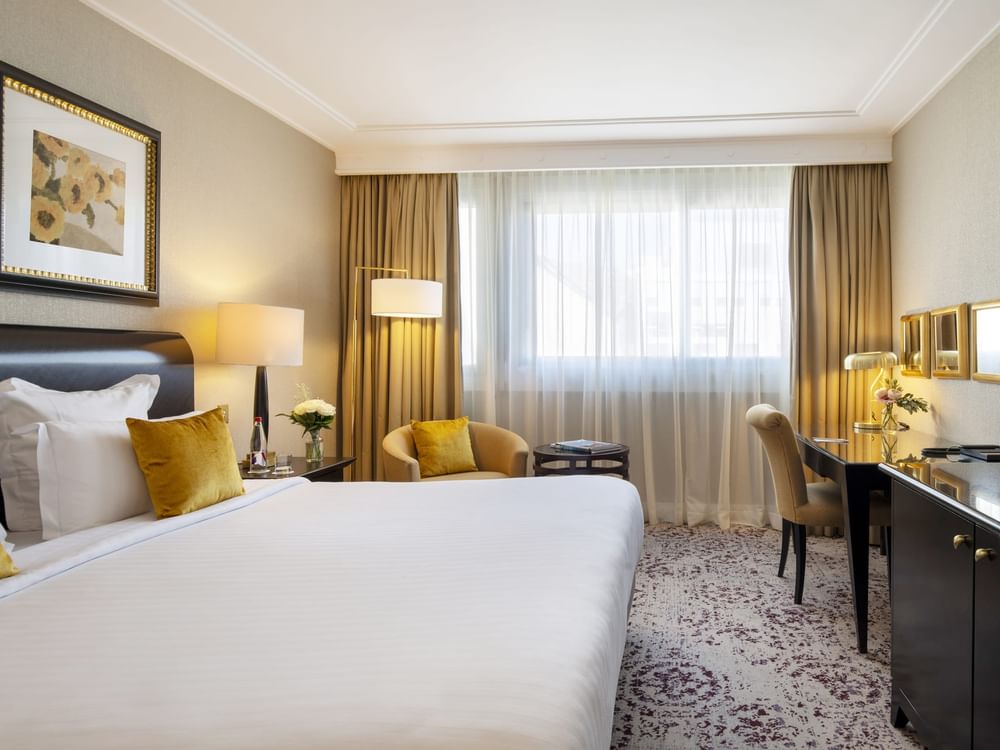 Tan armchair and desk by a white bed under a floor lamp near a window in The Montaigne Suite at Warwick Paris Champs Elysées