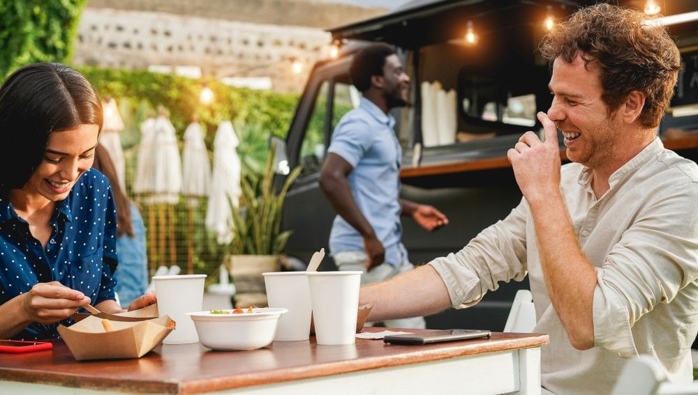 A man and woman sit at a table beside a food truck, laughing as they eat.