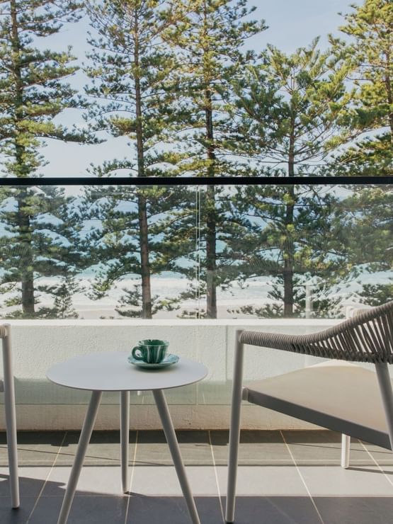 Ocean View King at Elysium Manly with a balcony table and green teacup by woven chairs under pine trees near a glass railing