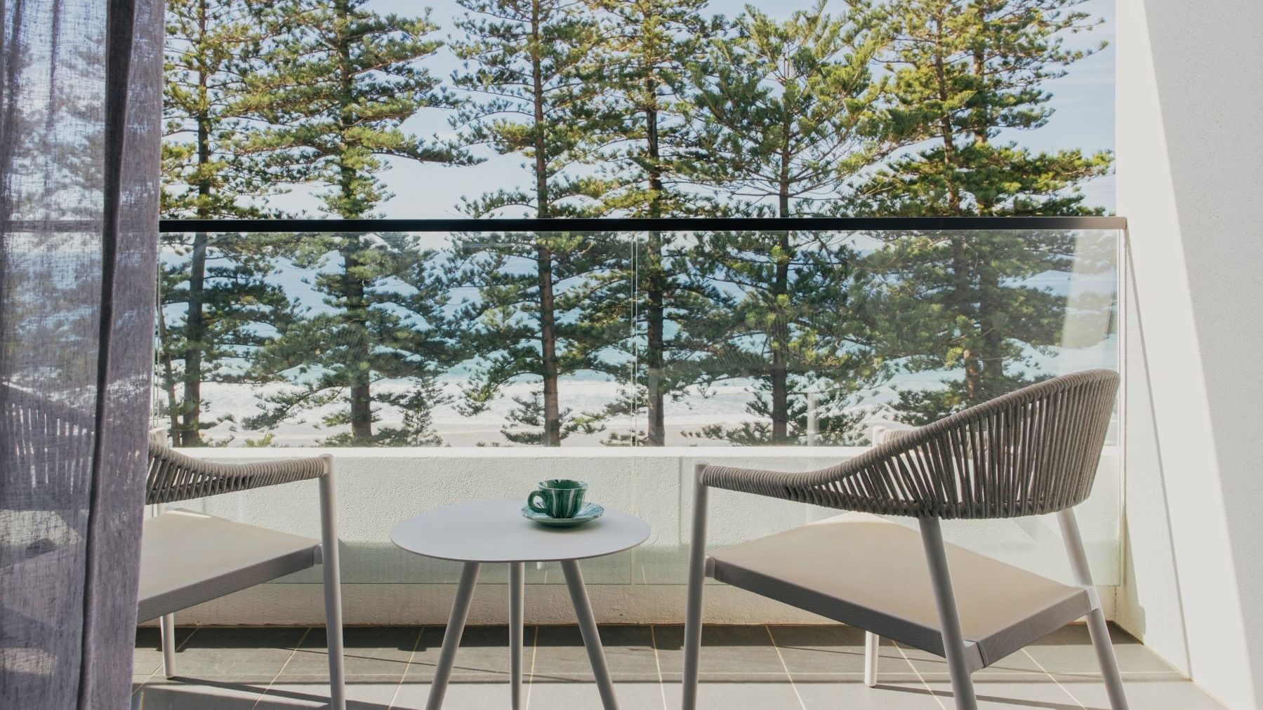 Ocean View King at Elysium Manly with a balcony table and green teacup by woven chairs under pine trees near a glass railing