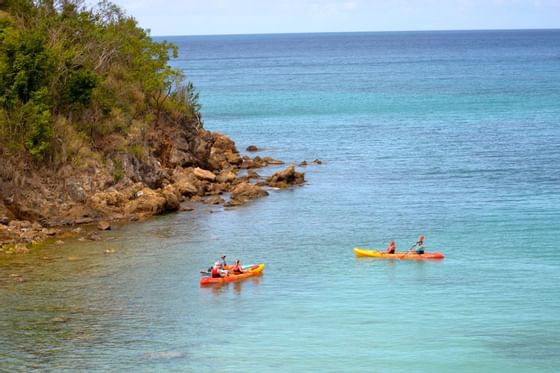 People paddling orange kayaks in calm turquoise ocean waters next to a rocky green cliff near Emerald Beach Resort