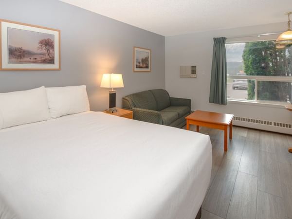 A room with a large bed, couch, coffee table, framed pictures, window, and a table with a kettle.