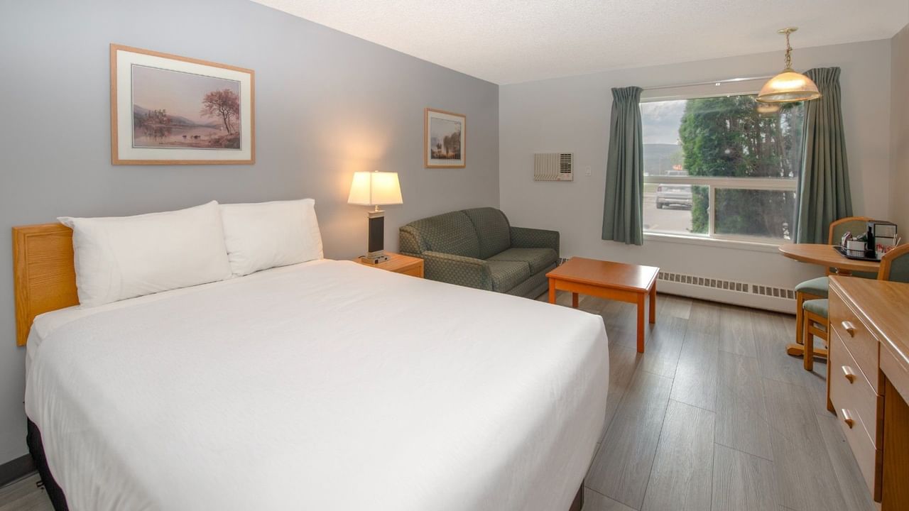 A room with a large bed, couch, coffee table, framed pictures, window, and a table with a kettle.