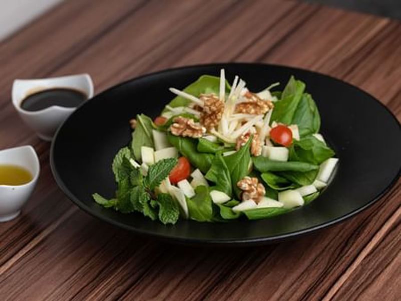 Spinach salad with walnuts and apples in a black bowl by two small cups of dressing in KVNT at Warwick Al Jubail