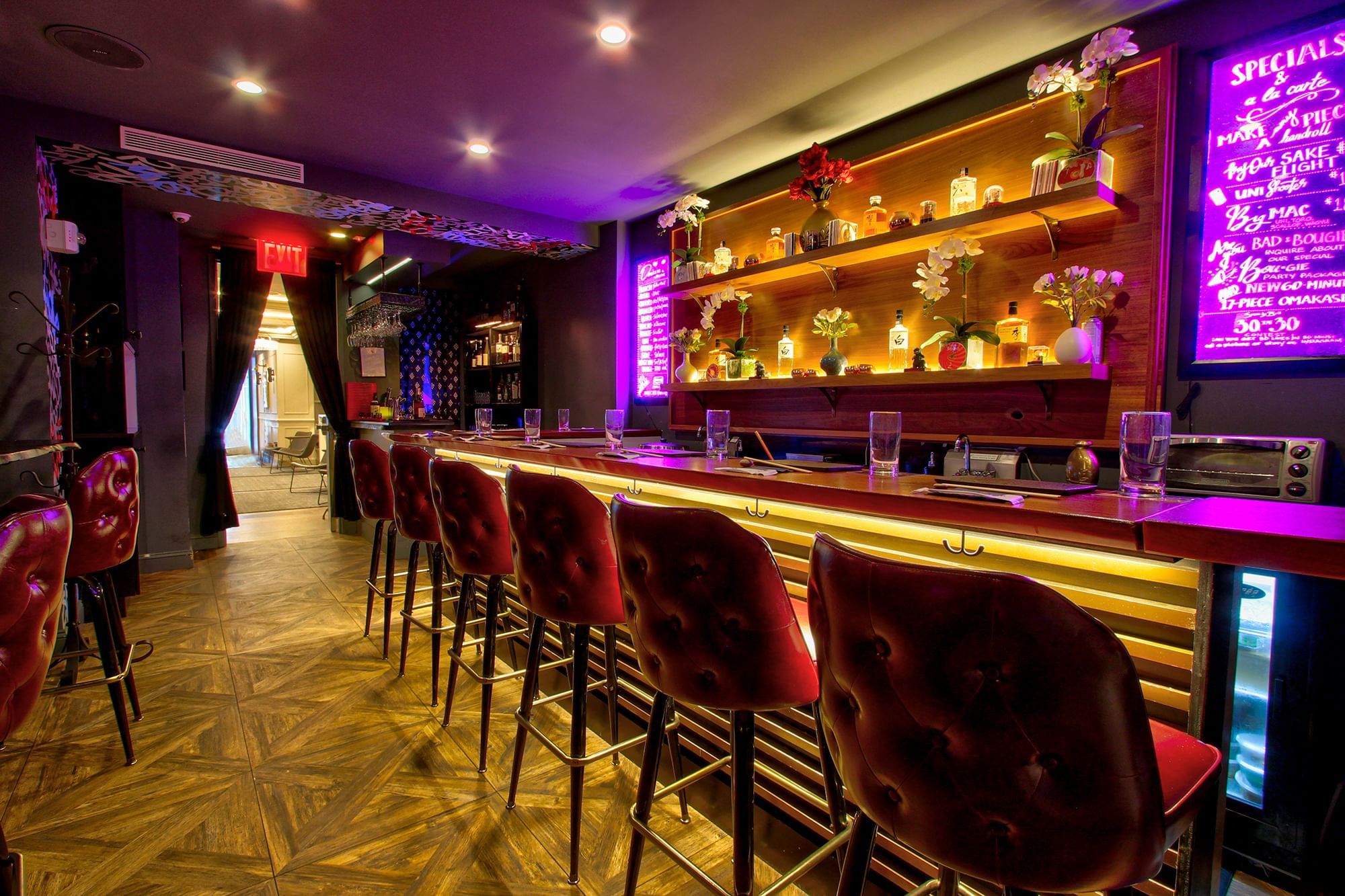 A dimly lit bar with a long counter, red leather stools, and shelves with decorative items.