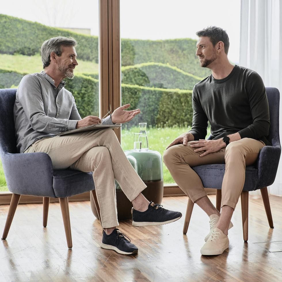 Two men in chairs converse in a room with a glass wall showing outdoor greenery.