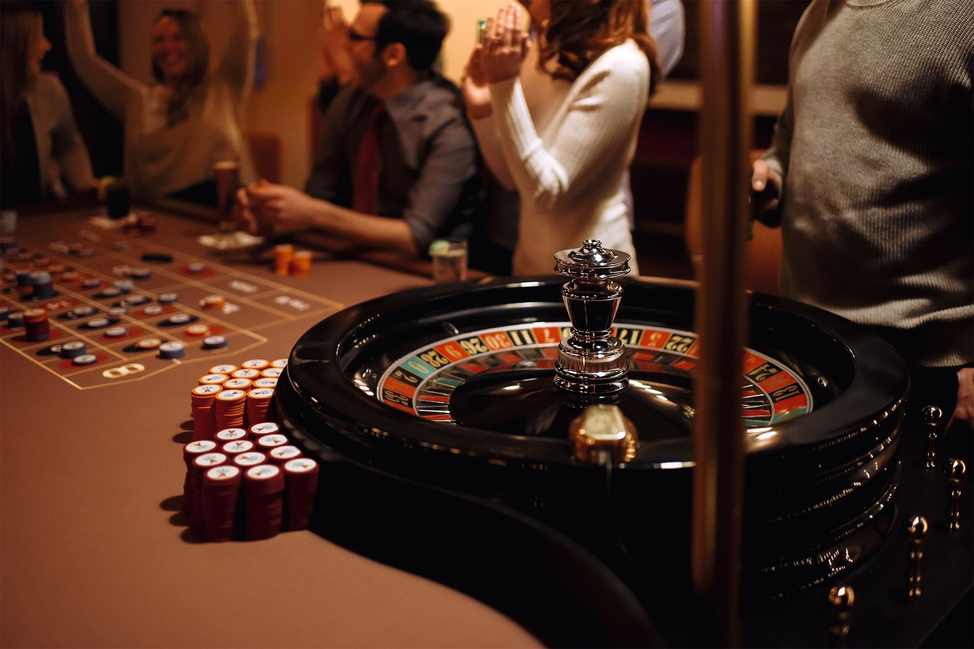 Roulette wheel by stacked casino chips as guests gather around a gaming table under warm pendant lights at The Brook