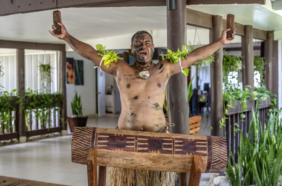 Traditional performer playing a carved wood drum by an open-air pavilion under the roof at Tambua Sands Beach Resort