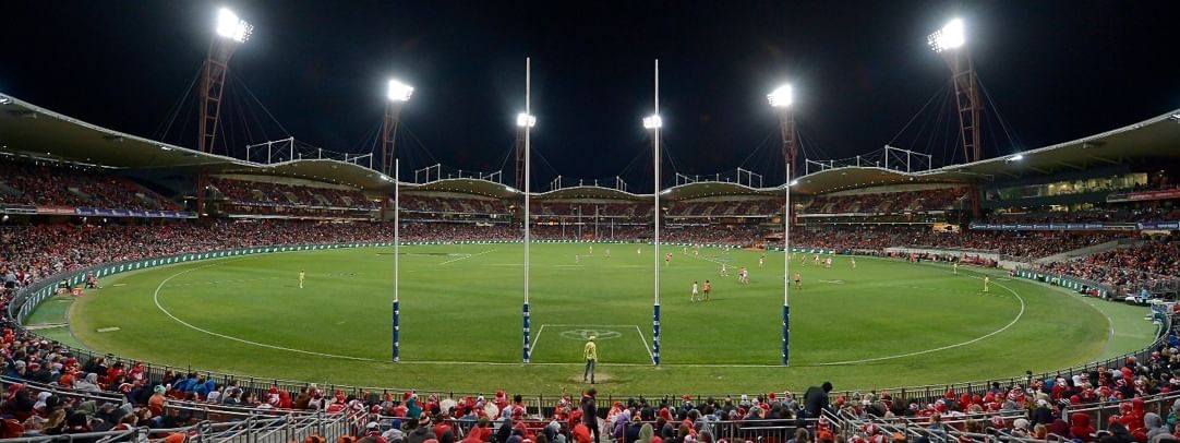 A large crowd is watching a night rugby match in a stadium with bright lights and a green field.
