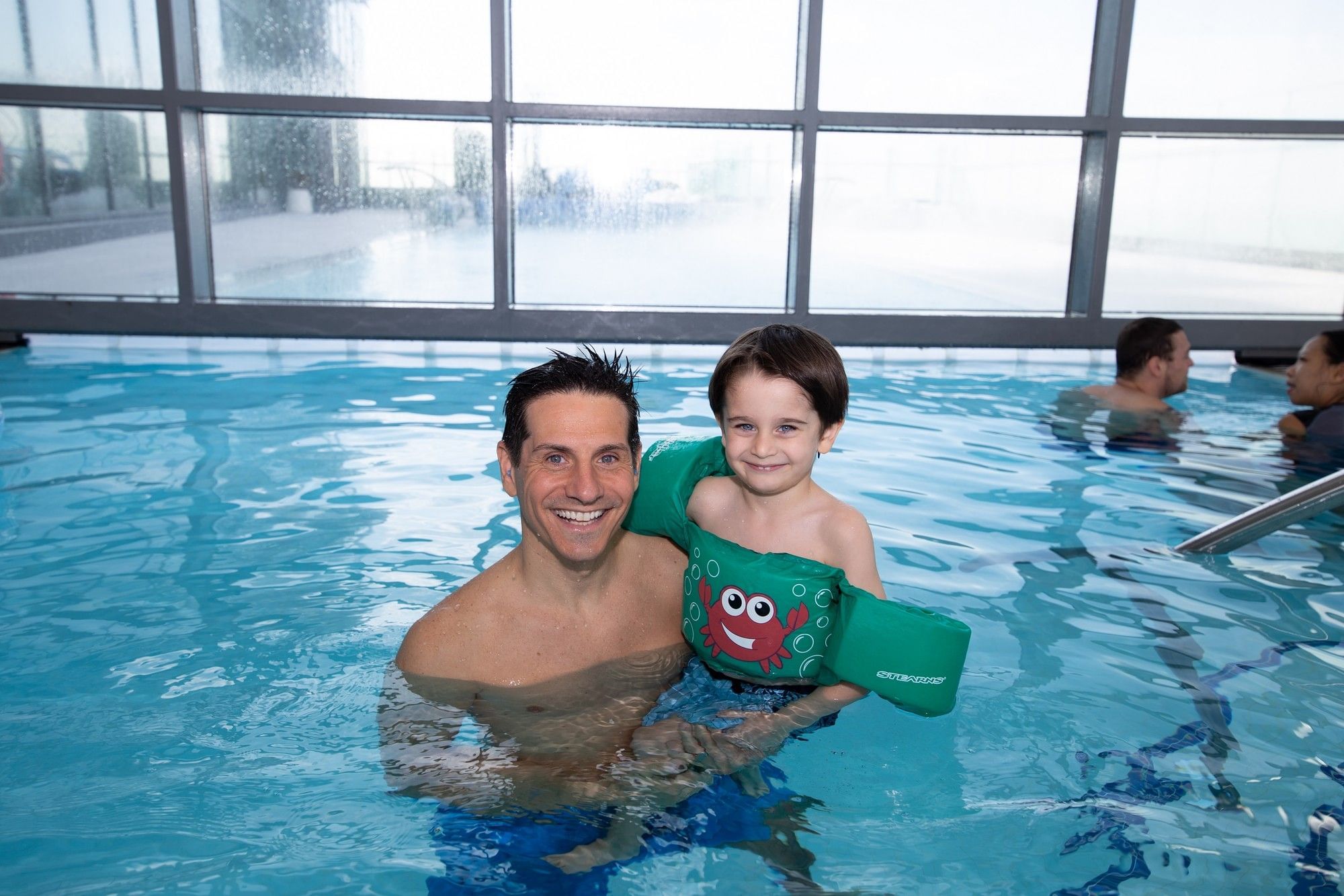 Father and chlid in a green crab floatie smiling inside a bright indoor swimming pool at Hotel X Toronto