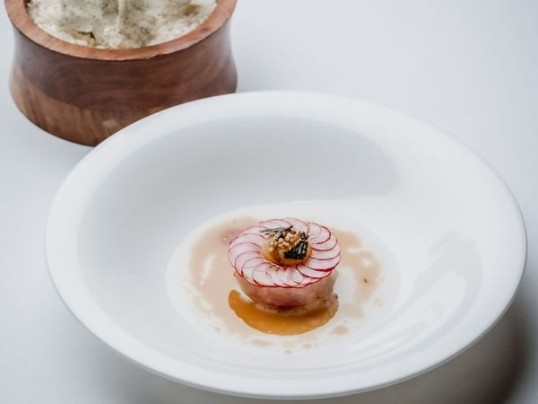 A white plate with a dish topped with radish slices and a small wooden bowl with garnish.