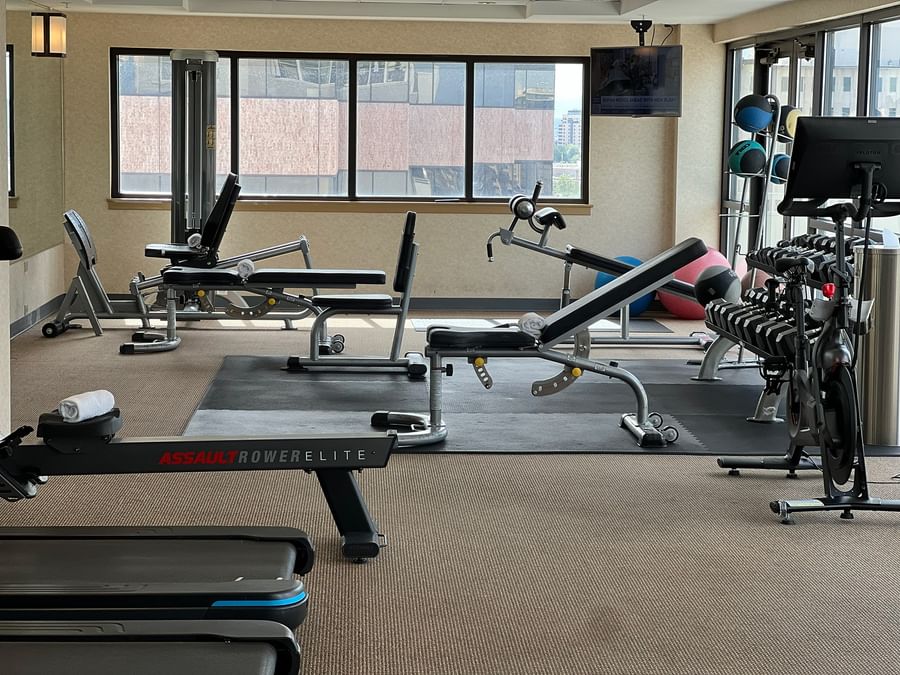 Gym weight benches by a rowing machine under a flat screen near windows at Warwick Denver