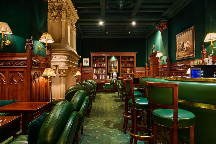 Green leather chairs arranged by wooden tables in bar with bookshelves and a fireplace at Hotel Westminster Paris