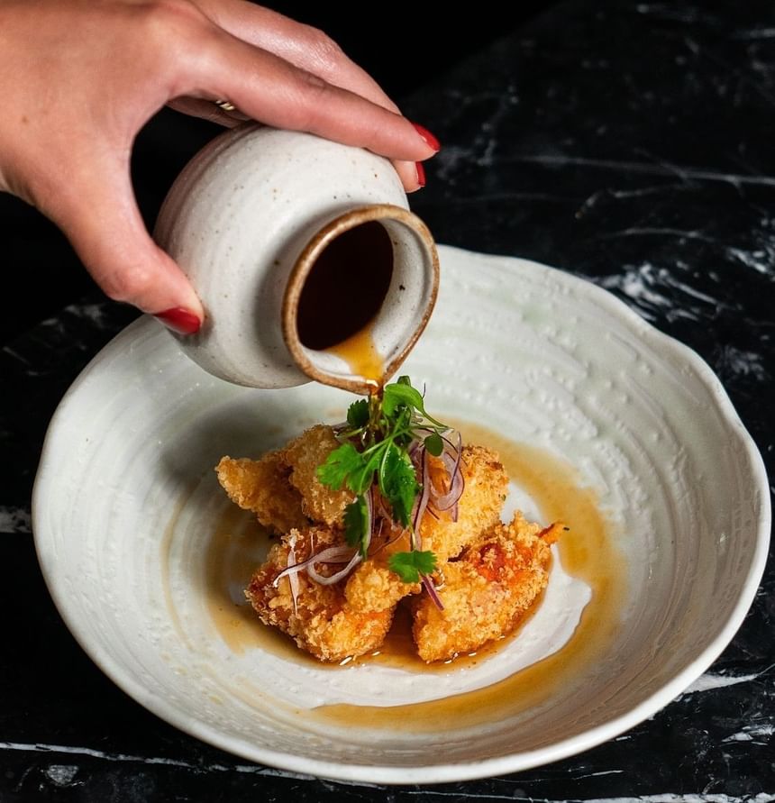 A hand pours sauce over crispy fried food with herbs on a white plate at The Kitchens