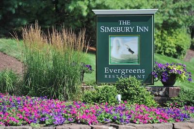 Green entrance sign for Evergreens Restaurant surrounded by vibrant pink and purple flowers outside at The Simsbury Inn