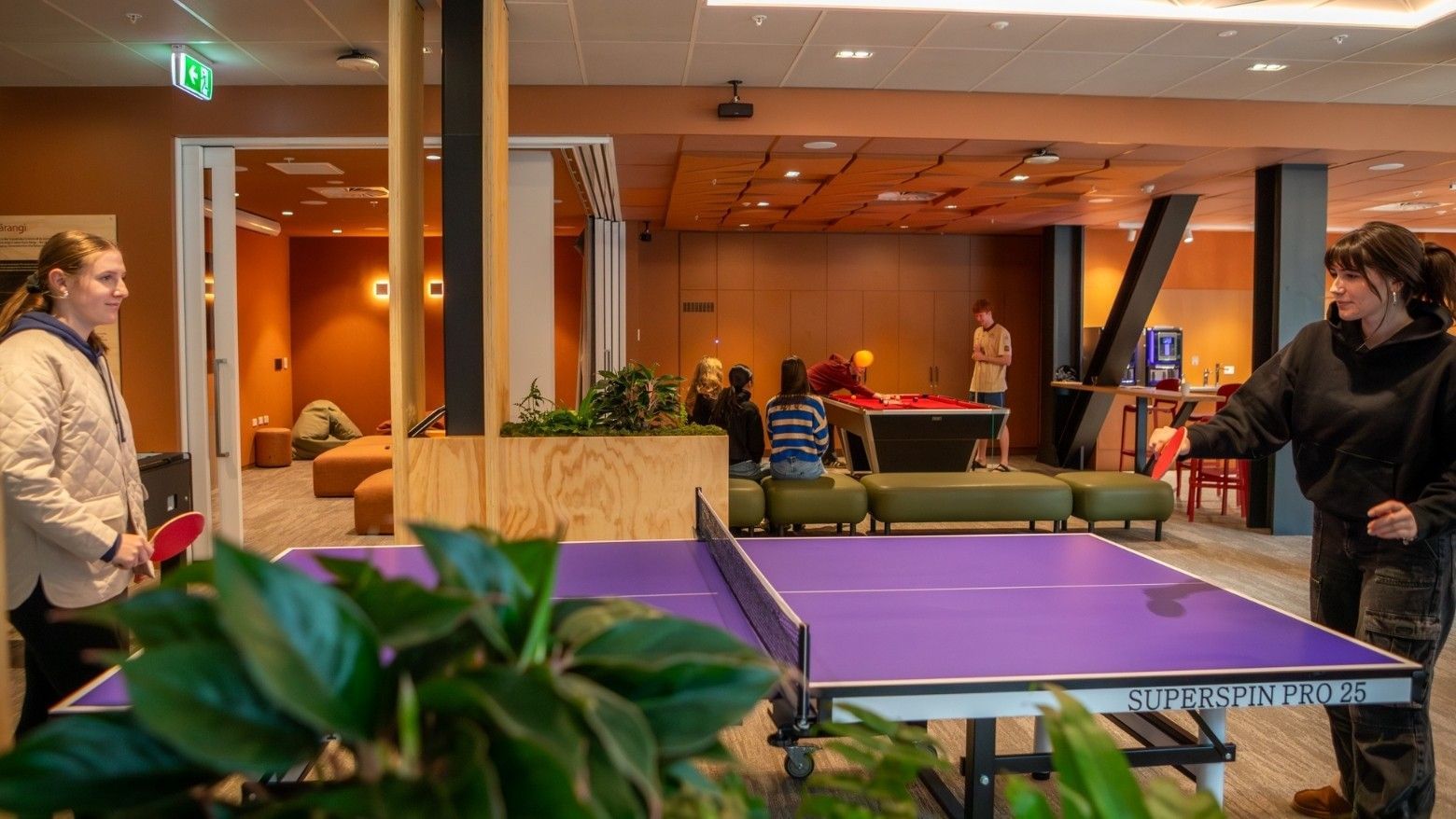 Modern recreation lounge with ping pong table, pool table, and guests enjoying games in warm-toned interior at Tupuārangi