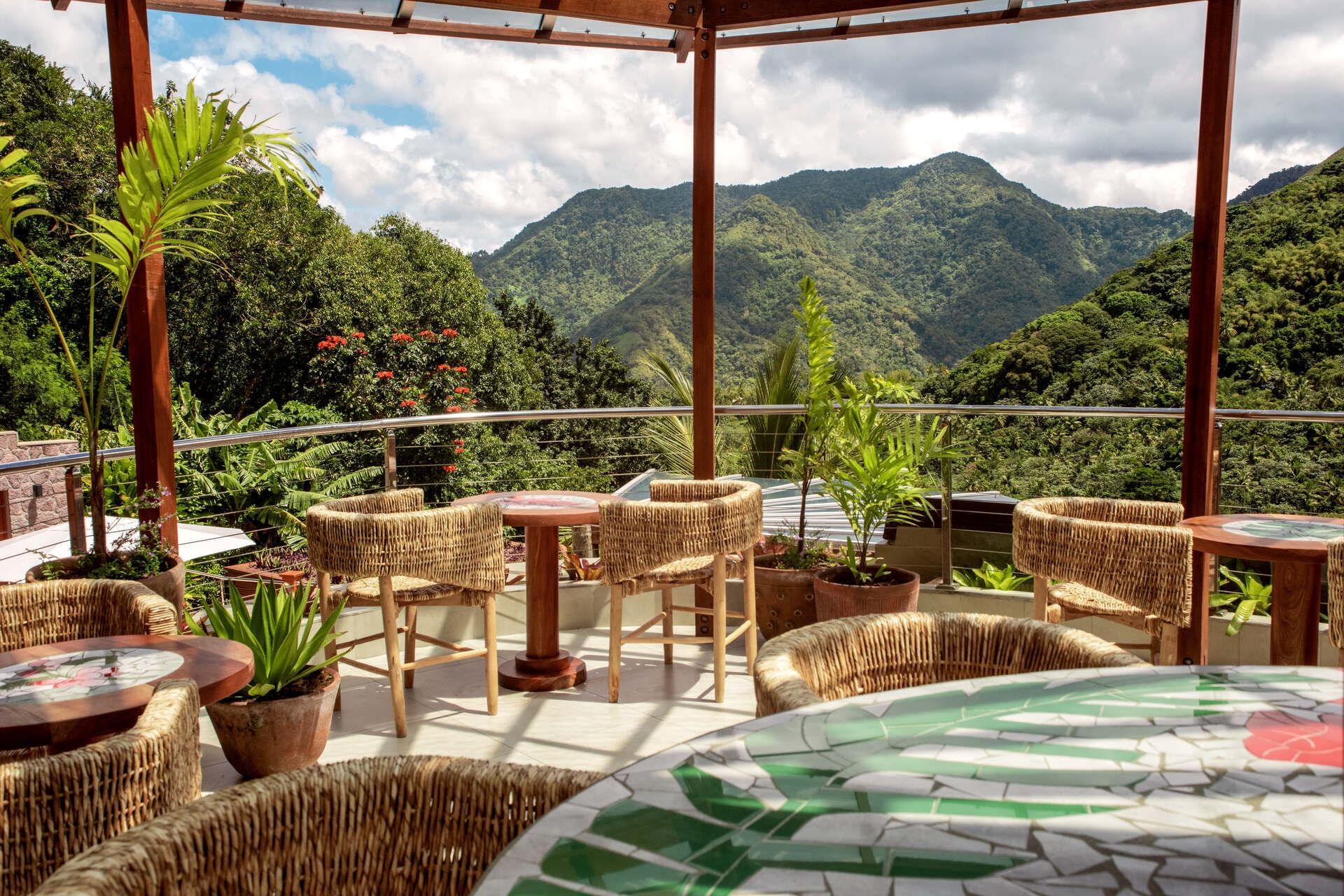 Outdoor patio with wicker chairs and tables, lush greenery, and scenic mountain view in the background.