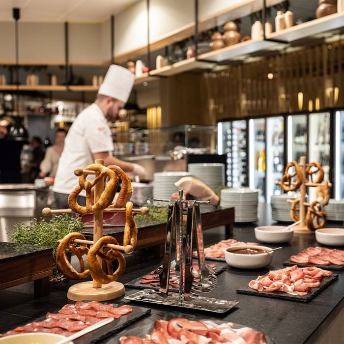 Koch in weißer Kochmütze bereitet Gerichte hinter einer Theke mit Wurstplatten und Brezeln zu.