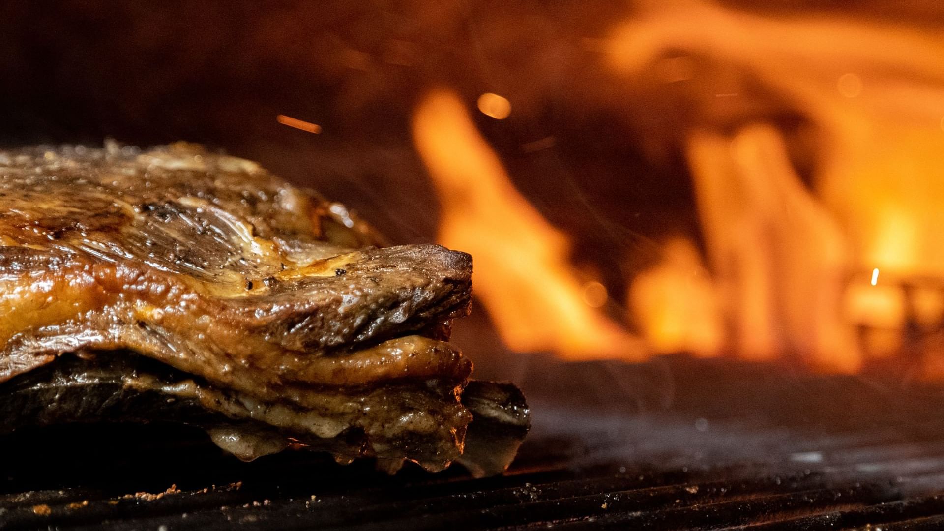 Primer plano de carne chisporroteando en una parrilla con llamas borrosas en De Cortez Grill House en Hacienda Del Mar Los Cabos