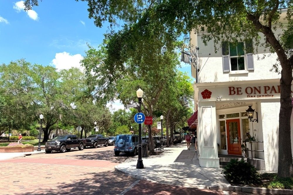 A cobblestone street beside a storefront with a red door, lined in mature trees. 